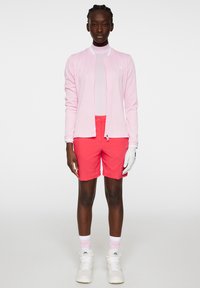 Ljusrosa texturerad kardiagan, vit långärmad topp, klara rosa shorts, vita sportskor och en vit handske. Enkelt design och livliga färger.