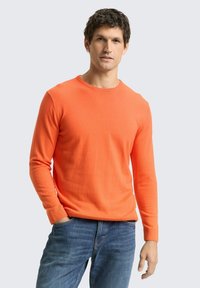 Fel oranje gebreide trui met lange mouwen en een ronde hals, gecombineerd met blauwe spijkerbroek. Soepele textuur en een aansluitend ontwerp.