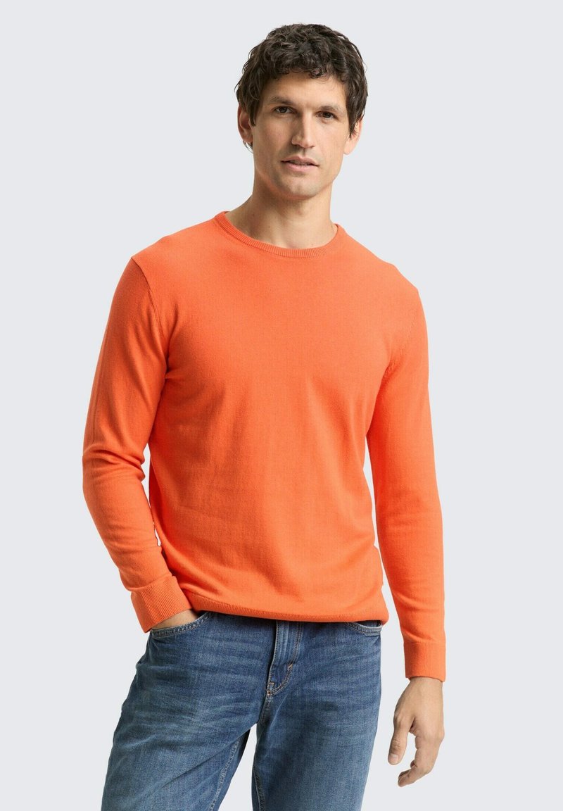 Fel oranje gebreide trui met lange mouwen en een ronde hals, gecombineerd met blauwe spijkerbroek. Soepele textuur en een aansluitend ontwerp.