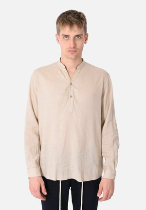 Giovane uomo con capelli corti che indossa una camicia beige a maniche lunghe con colletto henley abbottonato, in piedi davanti a uno sfondo bianco uniforme.