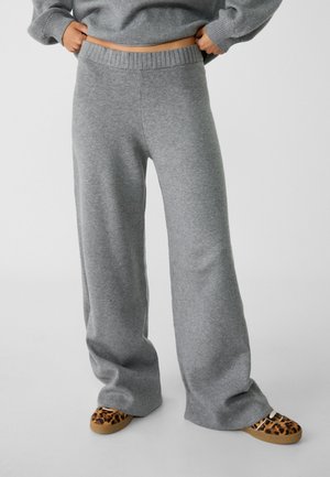 Personne portant un pantalon large en maille gris et un pull assorti, tenant la taille avec les mains, chaussures slip-on à motif léopard visibles.
