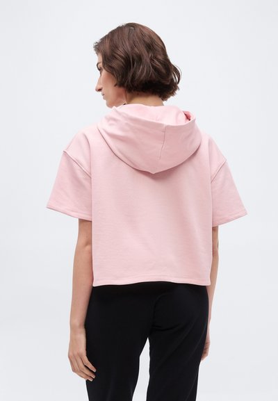 Woman with short brown hair wearing a light pink short-sleeve hooded top and black pants, facing away against a plain background.