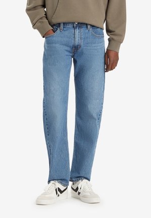 Homme portant un jean droit bleu clair, un sweat-shirt beige et des baskets blanches avec des détails noirs, debout avec une main dans la poche.