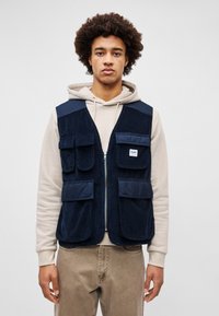 Gilet di velluto a righe blu navy con chiusura a zip, dotato di quattro tasche frontali. Indossato sopra un hoodie beige chiaro, con una vestibilità rilassata e tessuto testurizzato.