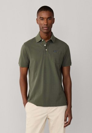 Hombre que lleva una camiseta tipo polo de manga corta verde oliva con botones y pantalones beige, de pie frente a un fondo liso gris claro.