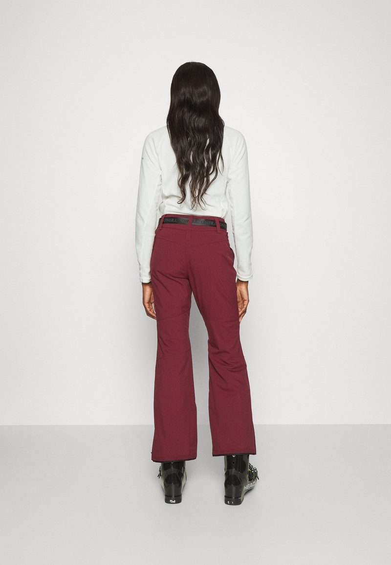 Burgundowe spodnie narciarskie mają rozszerzany krój oraz czarny pas z logo. Materiał wydaje się być wytrzymały, odpowiedni do sportów zimowych, o gładkiej teksturze.