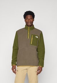 Pullover in pile verde e marrone, caratterizzato da un colletto alto, tasca con zip e accenti gialli a contrasto lungo la zip e l'orlo.