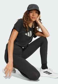 Crni kratki top s reflektivnim logotipom, crne tajice i bijele atletske cipele s crnim detaljima; model nosi crnu kapu s glatkom teksturom.