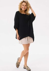 Czarny, wełniany, oversize sweter z rękawami w kształcie skrzydeł nietoperza oraz prążkowanym wykończeniem, noszony na jasnobeżowych szortach. Dopełnieniem stylizacji są buty w wężowy wzór.