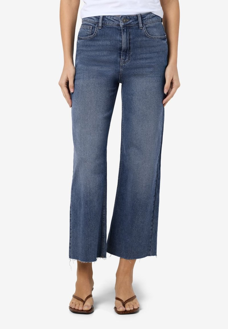 Γυναίκα που φοράει καμπάνα σιλουέτα μπλε τζιν με ραμένες άκρες και καφέ επίπεδες σάνταλ, που στέκεται μπροστά σε λευκό φόντο.