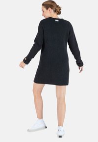 Robe pull noire en maille, longueur mi-cuisse, avec des manches longues, accent de boutons sur les poignets, texture côtelée et décolleté rond.
