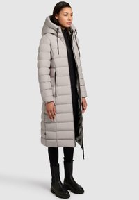 Lys grå lang pufferjakke med hætte, der har vandrette quiltede mønstre, sideskaber og en skinnende inderside, parret med sorte støvler og leggings.