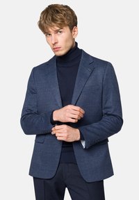 Niebieska marynarka w kratę z wyraźnymi ramionami, zapięciem na jeden guzik oraz klapami w kształcie notcha, połączona z ciemnym golfem pod spodem.