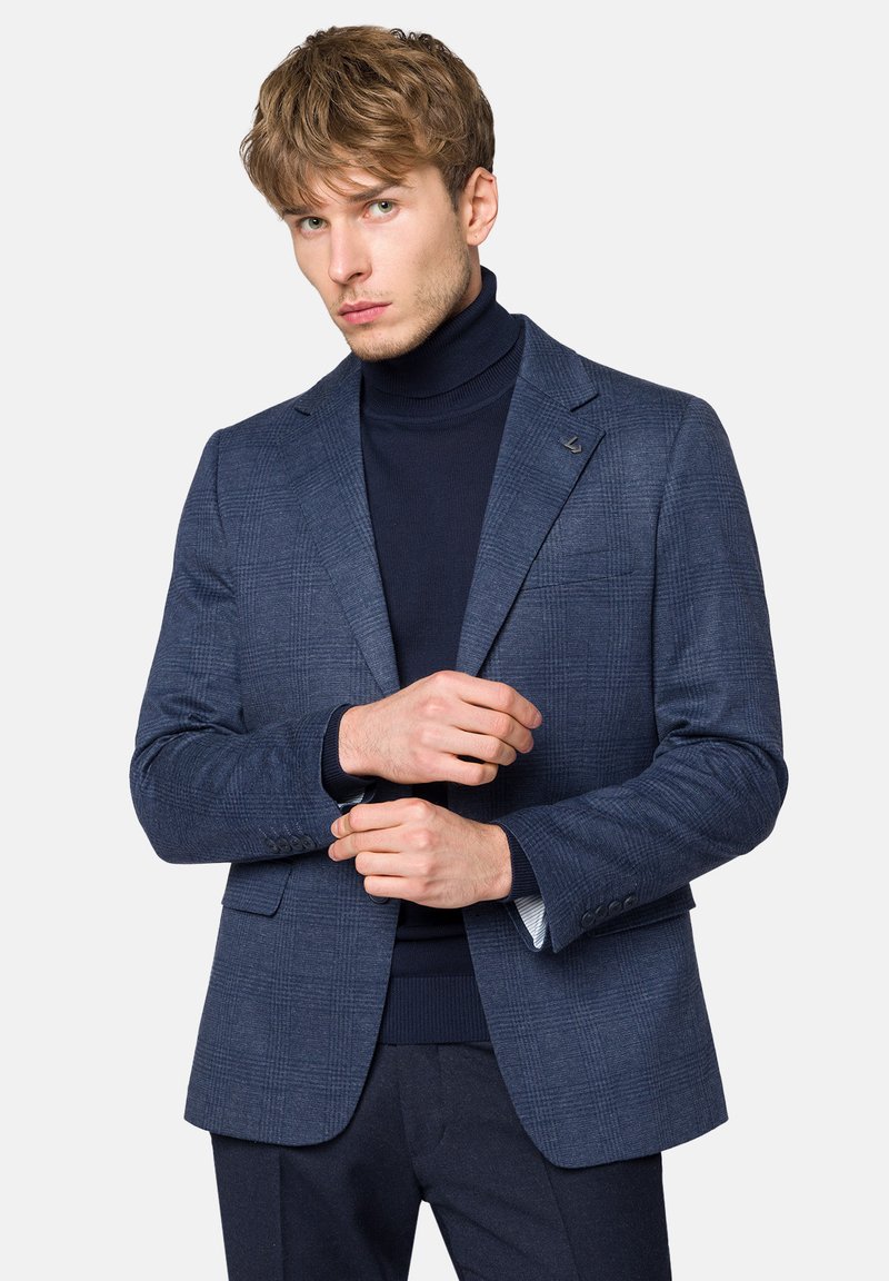 Niebieska marynarka w kratę z wyraźnymi ramionami, zapięciem na jeden guzik oraz klapami w kształcie notcha, połączona z ciemnym golfem pod spodem.