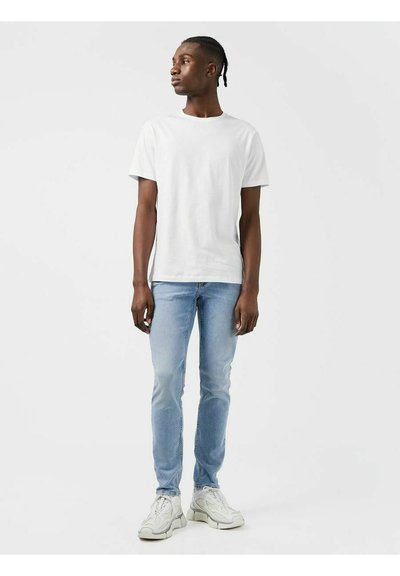 Witte katoen t-shirt, lichtblauwe slim-fit jeans en witte sneakers. Model staat voor een neutrale achtergrond, iets naar links gekeerd.