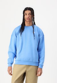 Puha anyagból készült kék pulóver, kerek nyakkivágással és bordás mandzsettával. Bézs nadrággal viselve, a mellkason egy kis logó található.