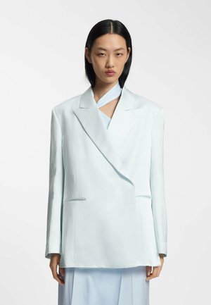 Femme aux cheveux noirs et raides portant un blazer bleu clair oversized et une jupe assortie sur un fond blanc uni.