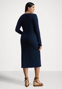 Vestido de malha canelada azul-marinho com mangas compridas, cintura ajustada e comprimento midi. Combinado com sandálias cunha castanhas. Textura suave com detalhes subtis.