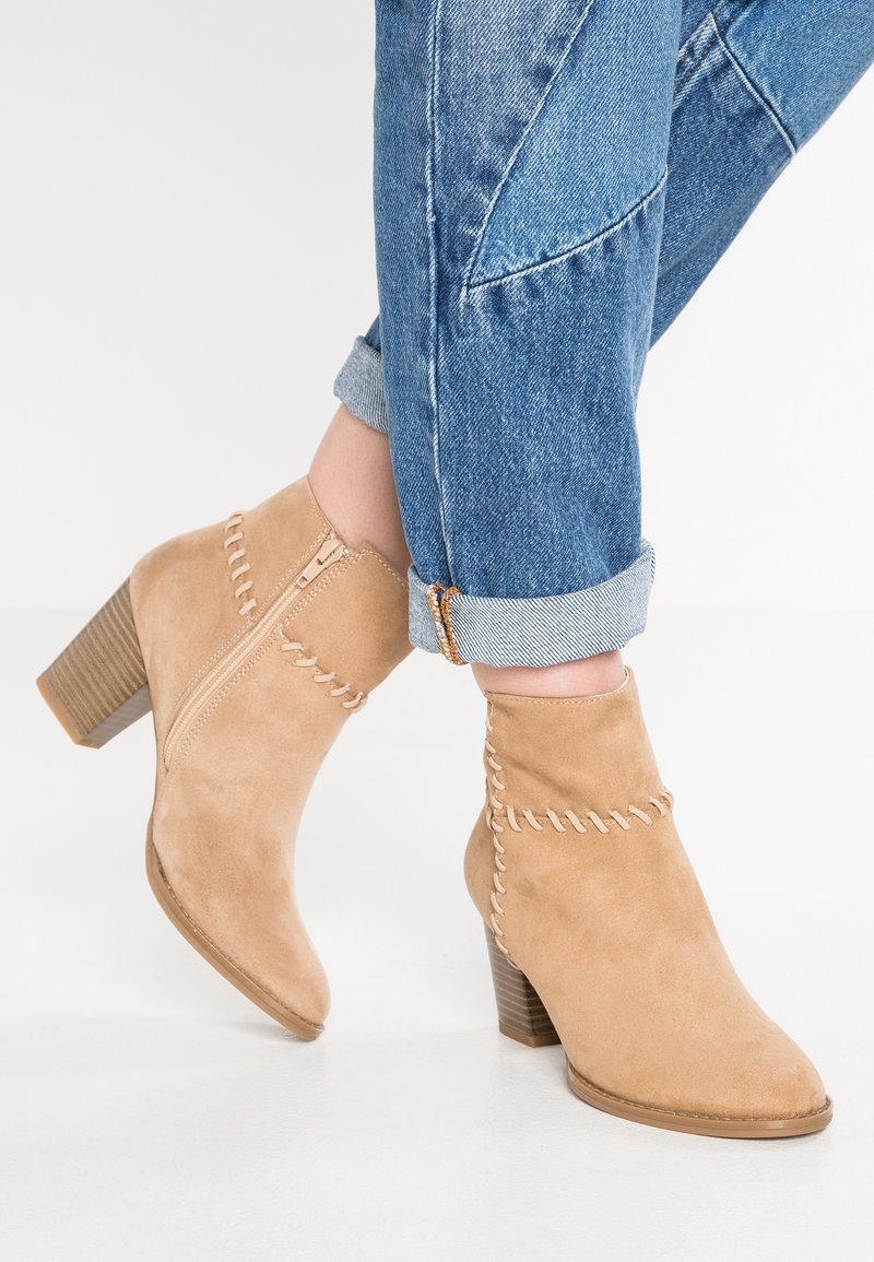 Bottes cheville en daim beige avec talons carrés et fermetures éclair latérales, portées avec un jean bleu retroussé sur un sol blanc.