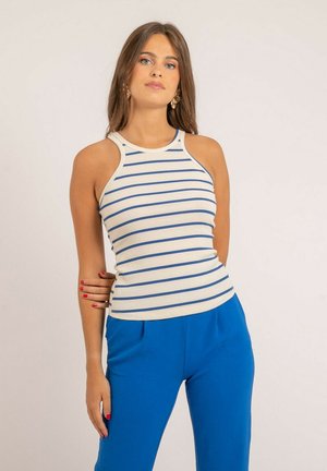 Jeune femme aux longs cheveux bruns portant un haut blanc sans manches à rayures bleues et un pantalon bleu vif, debout devant un fond uni.