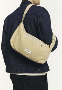 Sac en toile beige à l'épaule avec une forme arrondie, doté d'une poche zipée à l'avant et d'une étiquette en tissu. Porté par-dessus une veste sombre.