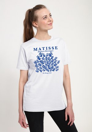 Kvinde med brunt hår sat op i hestehale, iført hvid T-shirt med blåt Matisse-snit og tekst, stående mod en lys baggrund og kiggende opad.