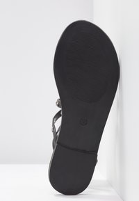Semelle noire d'une sandale plate avec grip texturé sur la zone de l'avant-pied, sangles visibles et petit logo près de la voûte plantaire sur fond blanc.