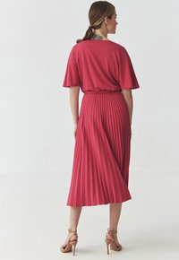 Abito rosso plissettato con maniche corte, caratterizzato da una texture liscia e una vita aderente. Indossato con tacchi con cinturino alla caviglia di color marrone chiaro.