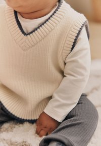 Knitted cream sleeveless sweater with ribbed pattern and navy trim, worn over a white long-sleeve shirt, paired with gray textured pants.