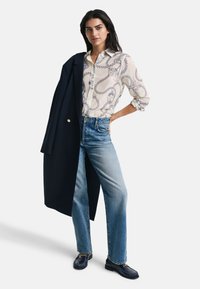 Langarm-Hemd mit cremigem Grund und dunkelblauem Seil-Print, kombiniert mit hellblauen geraden Jeans und einem dunklen Mantel. Schwarze Slipper sind inklusive.