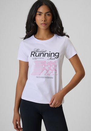 Kobieta z długimi ciemnymi włosami w białej koszulce z napisem "Afterlight Running Tour" i różowymi grafikami biegaczki, pozuje z jedną ręką na biodrze.