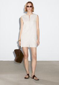 Beige sleeveless dress with a front slit, paired with a dark brown suede bag and flat sandals. Smooth texture and minimalistic design.