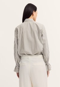 Gestreifte Bluse in Creme und Beige mit einem Kragen, elastischen Bündchen und einem lässigen Schnitt. Kombiniert mit hoch geschnittenen weißen Hosen.