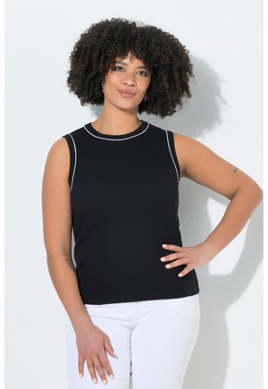 Femme aux cheveux bouclés portant un haut noir sans manches avec une bordure blanche et un pantalon blanc, debout avec une main sur la hanche.