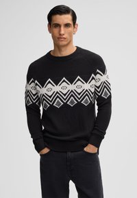 Zwarte gebreide trui met een geometric patroon in wit en grijs over de borst, met een ronde halslijn en geribbelde boorden en zoom.