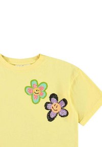 Gul bomulds t-shirt med to broderede blomsterdesigns, en i grøn og pink og en anden i lilla og sort, begge med smilende ansigt.