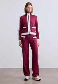 Μπορντό φόρμα με λευκές λεπτομέρειες· το μπουφάν με φερμουάρ διαθέτει δύο μπροστινές τσέπες, σε συνδυασμό με παντελόνι φαρδύ στα πόδια. Λεία, απαλή υφή υφάσματος.