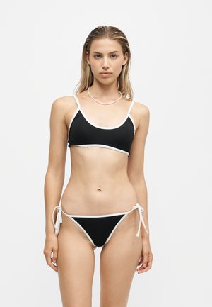 Jeune femme aux longs cheveux portant un bikini noir avec une bordure blanche, debout devant un fond gris clair uni.
