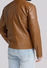 Giacca di pelle marrone con accenti trapuntati sulle spalle, vestibilità slim e dettagli con zip sulle maniche; abbinata a jeans blu chiaro.