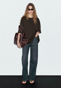 Maglione marrone oversize, jeans scuri a vita alta, e borsa lucida marrone. Il modello indossa occhiali da sole neri e sandali aperti.