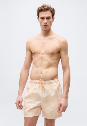 Young man with curly hair wearing pale orange and white vertical striped swim shorts, standing against a plain light background.