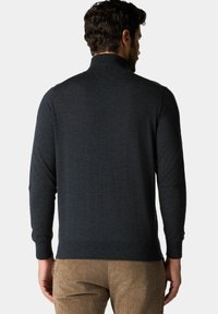 Uomo che indossa un maglione a maniche lunghe color carbone scuro e pantaloni di velluto a coste marrone chiaro, visto di spalle su uno sfondo semplice.