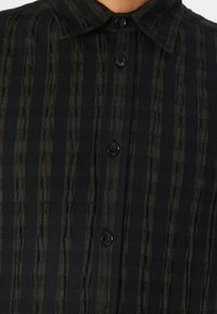 Camisa a rayas de color negro y verde oliva oscuro con un diseño de botones, presenta un cuello con solapa y una tela texturizada con patrones verticales.