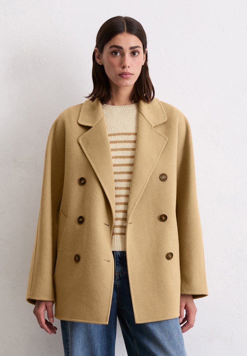 Double-breasted, beige wool coat with large brown buttons, notched lapel, and a relaxed fit, worn over a cream and brown striped sweater.