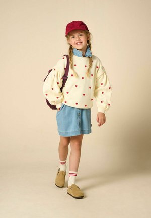 Fille souriante aux cheveux tressés portant une casquette rouge, un pull à motifs de cœurs, une jupe en jean et des sandales avec des chaussettes, portant un sac à dos.
