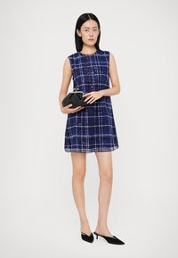 Robe sans manches bleu avec un motif à carreaux blancs, tissu texturé et jupe évasée ; associée à une pochette noire et des chaussures noires à talons pointus.