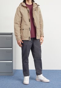 Beige huva jacka med justerbara dragsko, stora framfickor och svarta detaljer. Kombinerad med en vinröd skjorta och mörkgrå byxor.