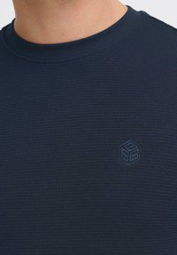 Tamsiai mėlynas tekstūruotas krūtinės apykaklės megztinis su subtiliu itin iškeltu logotipu ant krūtinės. Medžiaga atrodo minkšta ir tampri.