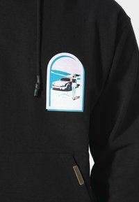 Sudadera negra con un gráfico azul y blanco de un coche vintage y una mujer con un sombrero de pie junto a él en la zona del pecho.