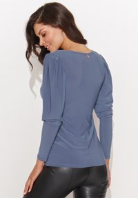 Niebieska bluzka z długim rękawem, z bufkami na ramionach i detalem w postaci dziurki na guzik z tyłu, w połączeniu z dopasowanymi czarnymi legginsami. Gładka tekstura materiału.